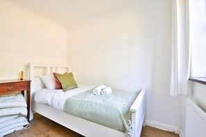 A single bed with a white headboard, a green and white striped blanket, and two rolled towels. There is a wooden nightstand with a lamp and a stack of white towels. - Homely 5 Bed House - 15 Mins to Cardiff City (Cardiff)