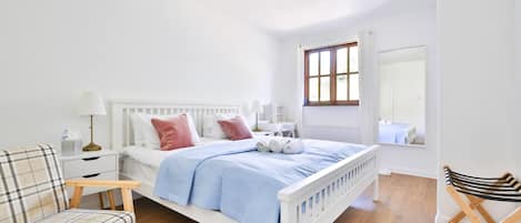 Une chambre à coucher avec un lit blanc, une chaise, une petite table et un miroir.