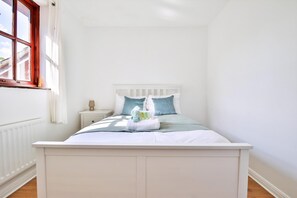 Una cama blanca con cabecera, una mesita de noche con lámpara y una ventana con vistas a edificios.
