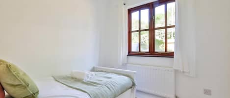 Uma cama de solteiro com colchão branco e roupa de cama verde, uma janela com cortinas brancas e um radiador na parede.