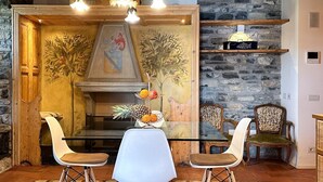 A dining area with a glass table, two chairs, a fruit centerpiece, and a stone wall with a mural. - Nestled on the hillside of Cernobbio, this enchanting 1608 structure, once a Convent, has undergone a meticulous restoration, now comprising three self-contained apartments. This unit with its tasteful décor, ensures a harmonious blend of style and comf (Cernobbio)