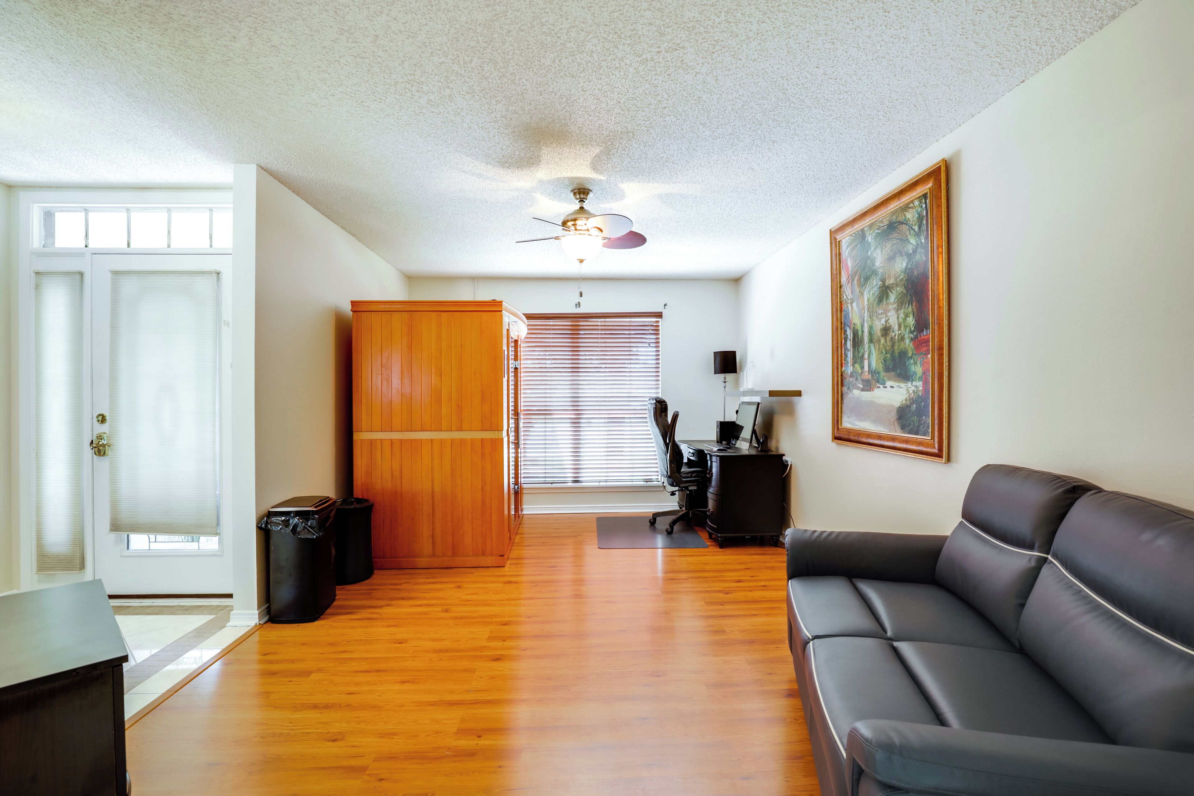 Sala de estar com piso de madeira, um sofá de couro preto, um armário de madeira, um ventilador de teto e uma pintura grande na parede.