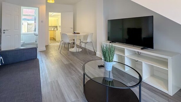 Ein modernes Wohnzimmer mit Flachbildfernseher, einem Glastisch und einem Essbereich mit weißen Stühlen.