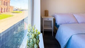 Ein Hotelzimmer mit Bett, Nachttisch und Fenster mit Blick auf ein Gebäude und eine Straße.