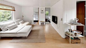 A modern, minimalist living room with a white sofa, a flat-screen TV mounted on the wall, and a wooden dining table.
