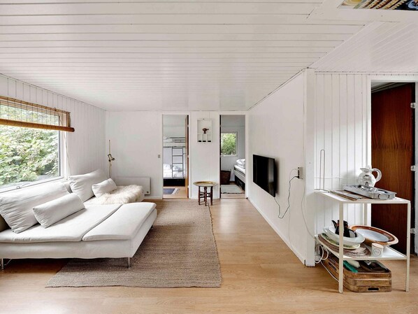 A modern, minimalist living room with a white sofa, a flat-screen TV mounted on the wall, and a wooden dining table.