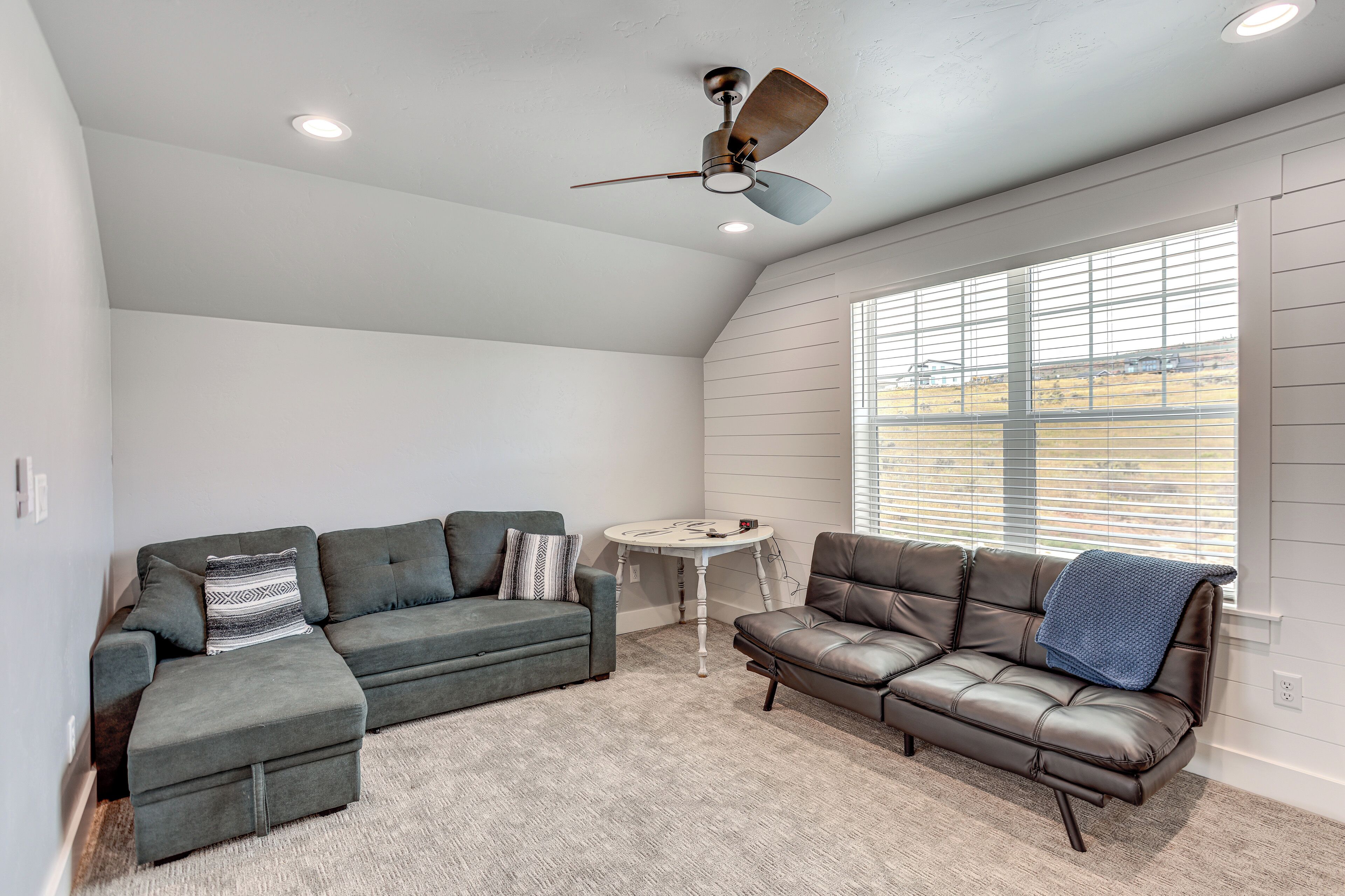 A room with two sofas, a ceiling fan, a small table, and a window with blinds.