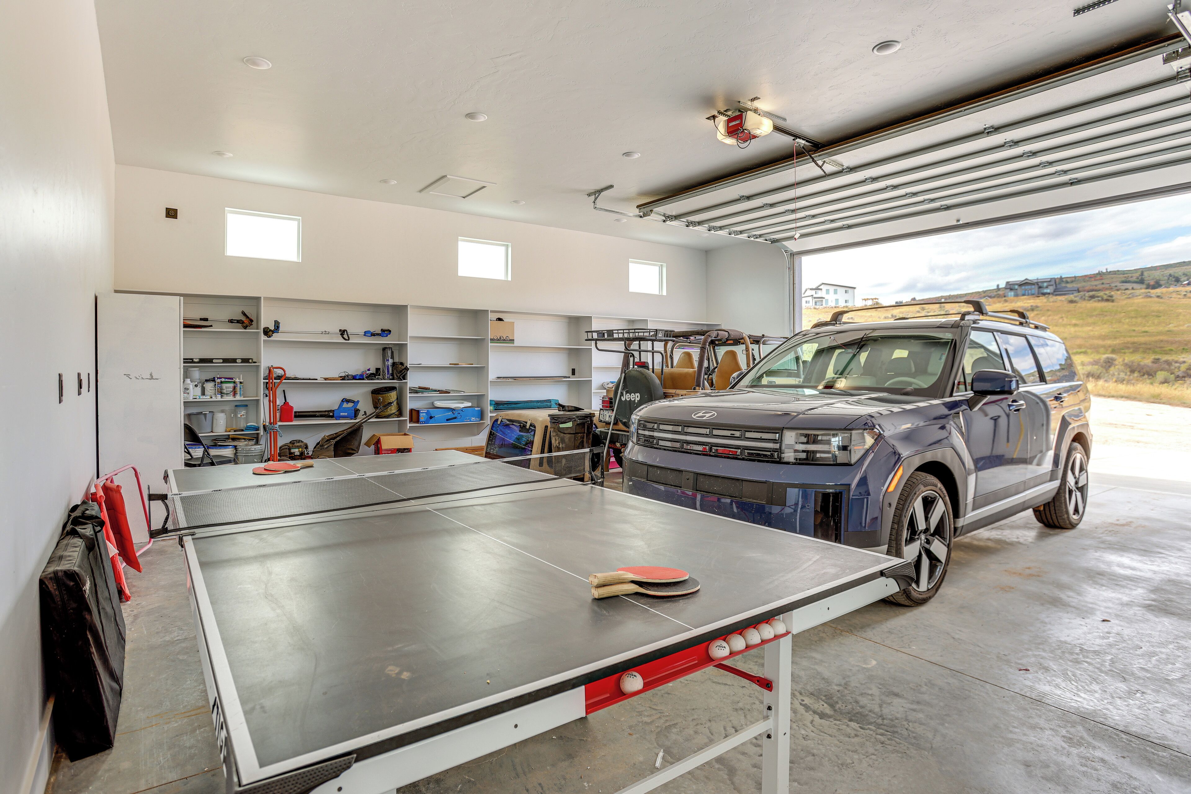A garage with a ping pong table, shelves with tools, and a Jeep SUV.