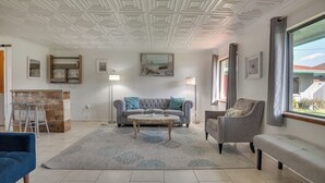 A living room with a grey sofa, a blue armchair, a coffee table, and a large window with curtains. - Beach Retreat w/ Pool Near Rocket Launches (Cape Canaveral)