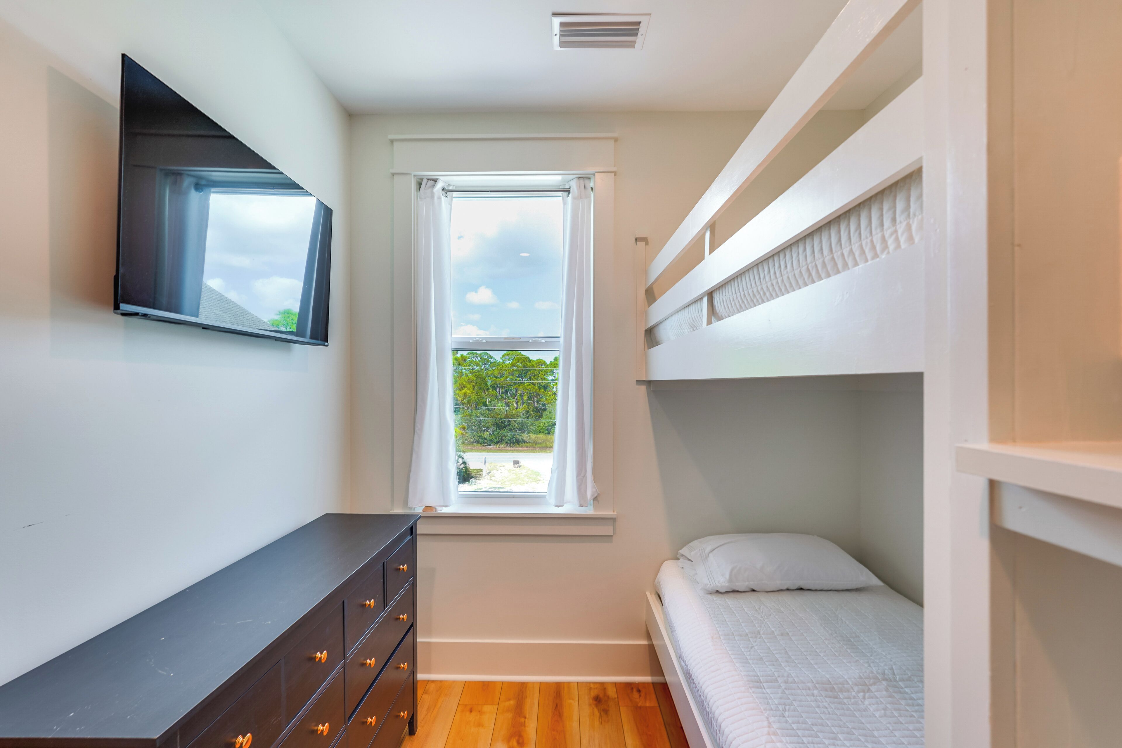 Un dormitorio pequeño con literas, un aparador con cajones, una ventana con cortinas y un televisor de pantalla plana montado en la pared.