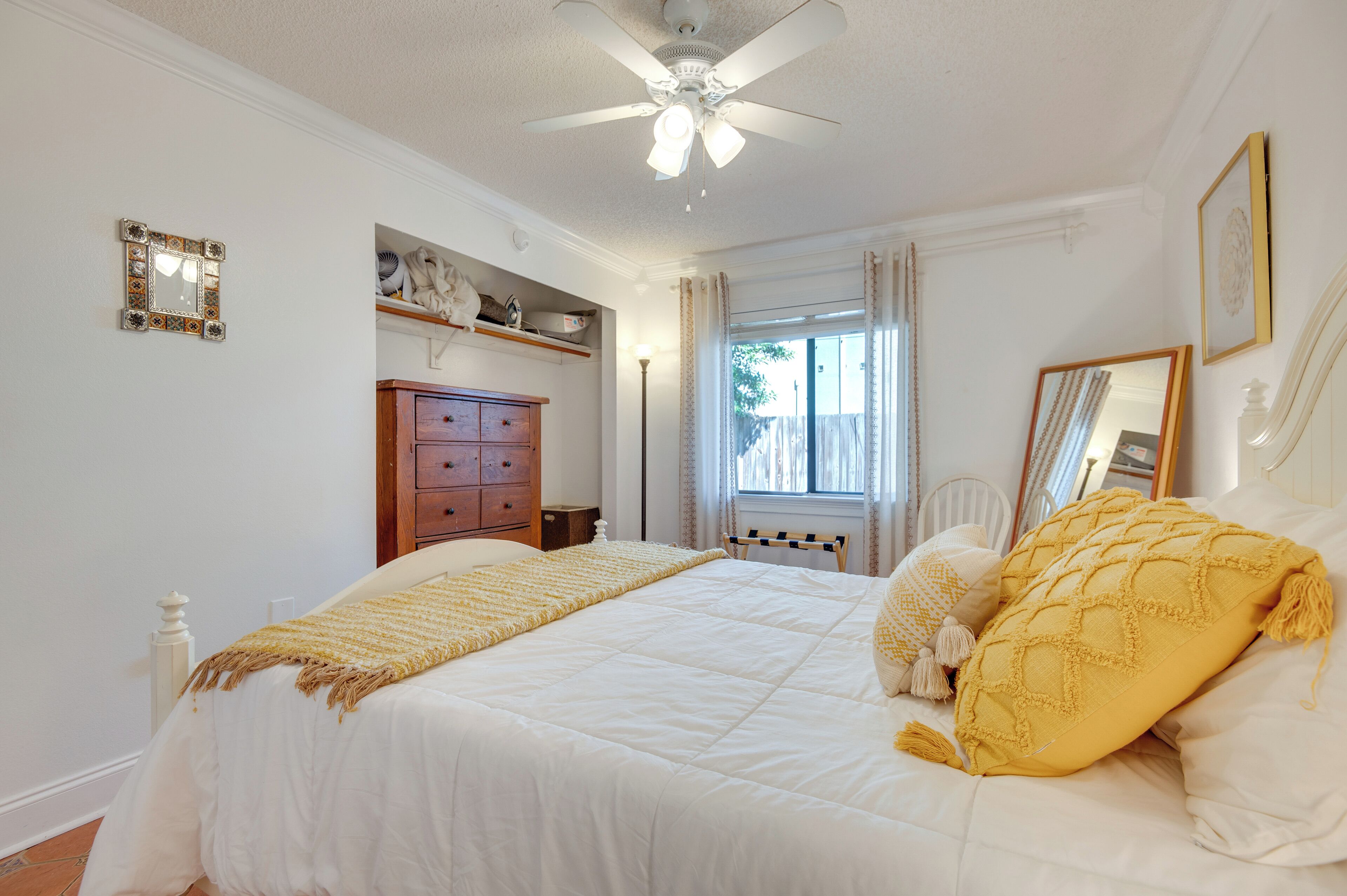 Een slaapkamer met een bed, een plafondventilator, een raam met gordijnen, een dressoir en een spiegel aan de muur.