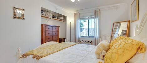 Een slaapkamer met een bed, een plafondventilator, een raam met gordijnen, een dressoir en een spiegel aan de muur.