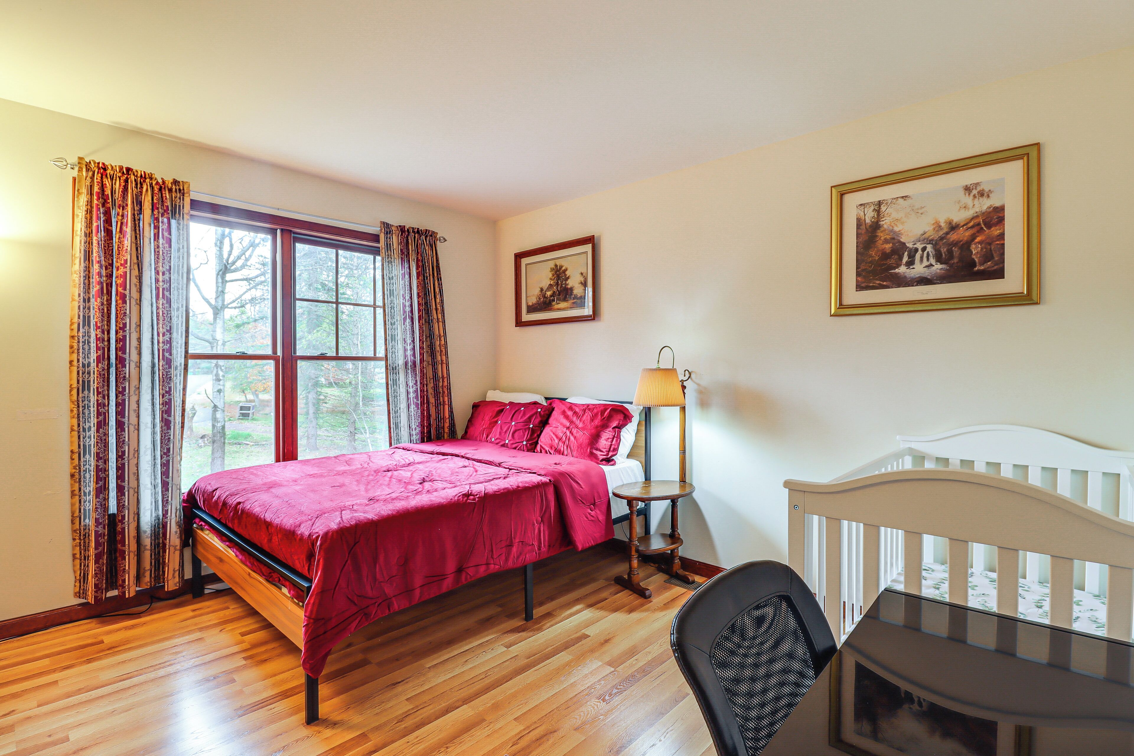 Une chambre avec un plancher en bois, un lit recouvert d ’une courtepointe rouge, une table de chevet avec une lampe, un berceau et deux tableaux encadrés accrochés au mur.