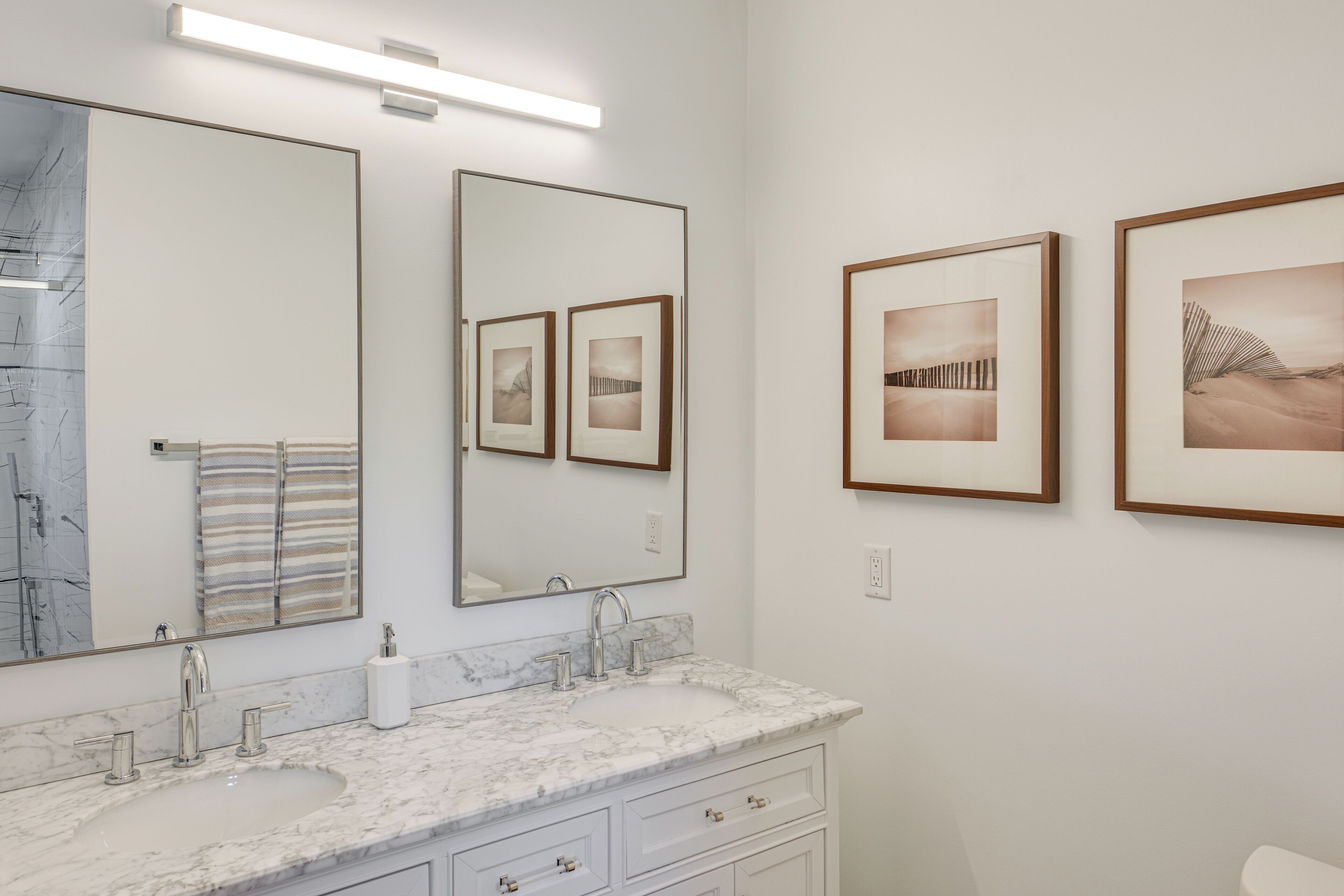 Un baño moderno con un lavamanos de mármol, dos llaves, un espejo grande y obras de arte enmarcadas en la pared.