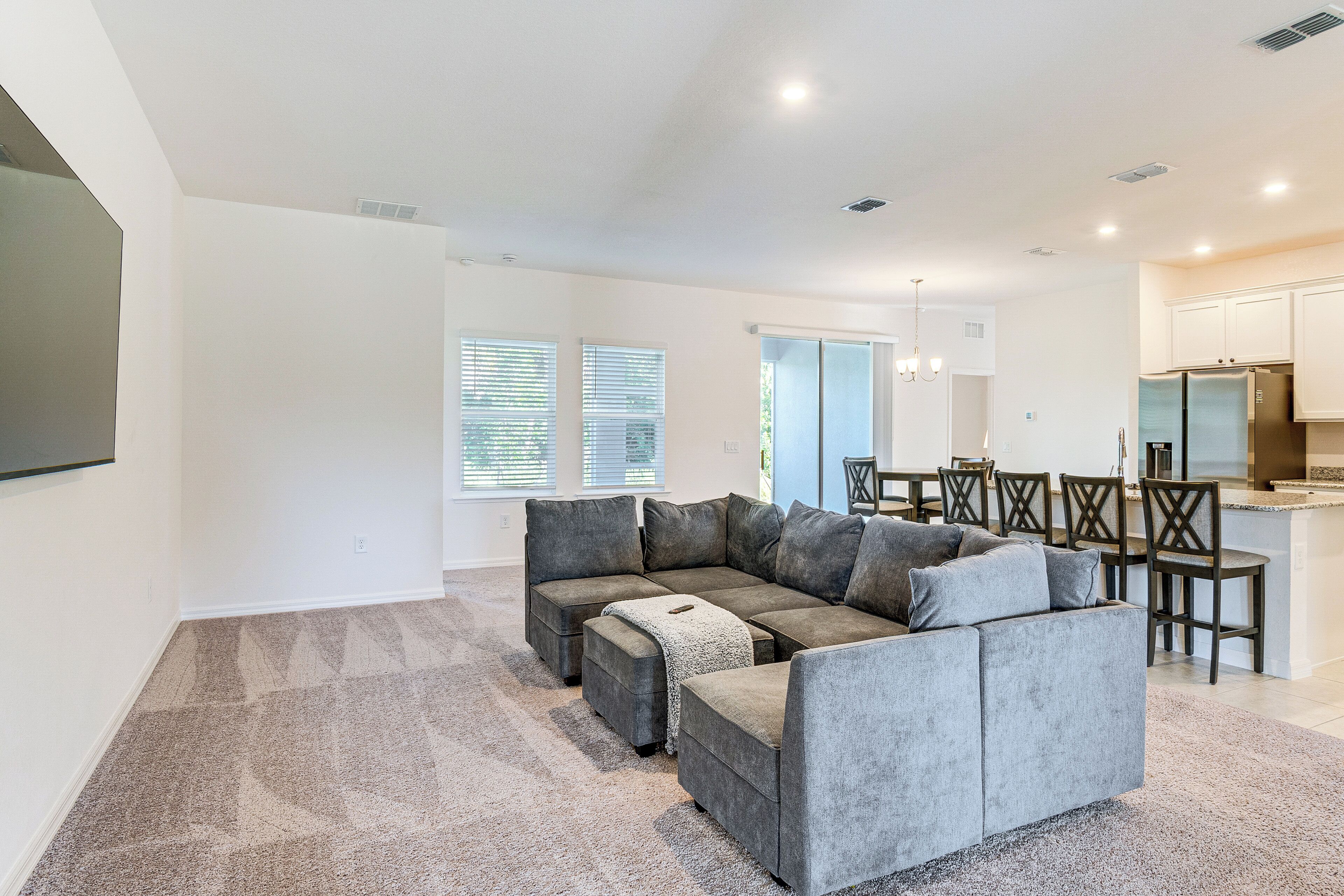 A modern living room with a sectional sofa, a dining area with a table and chairs, and a kitchen with a refrigerator.