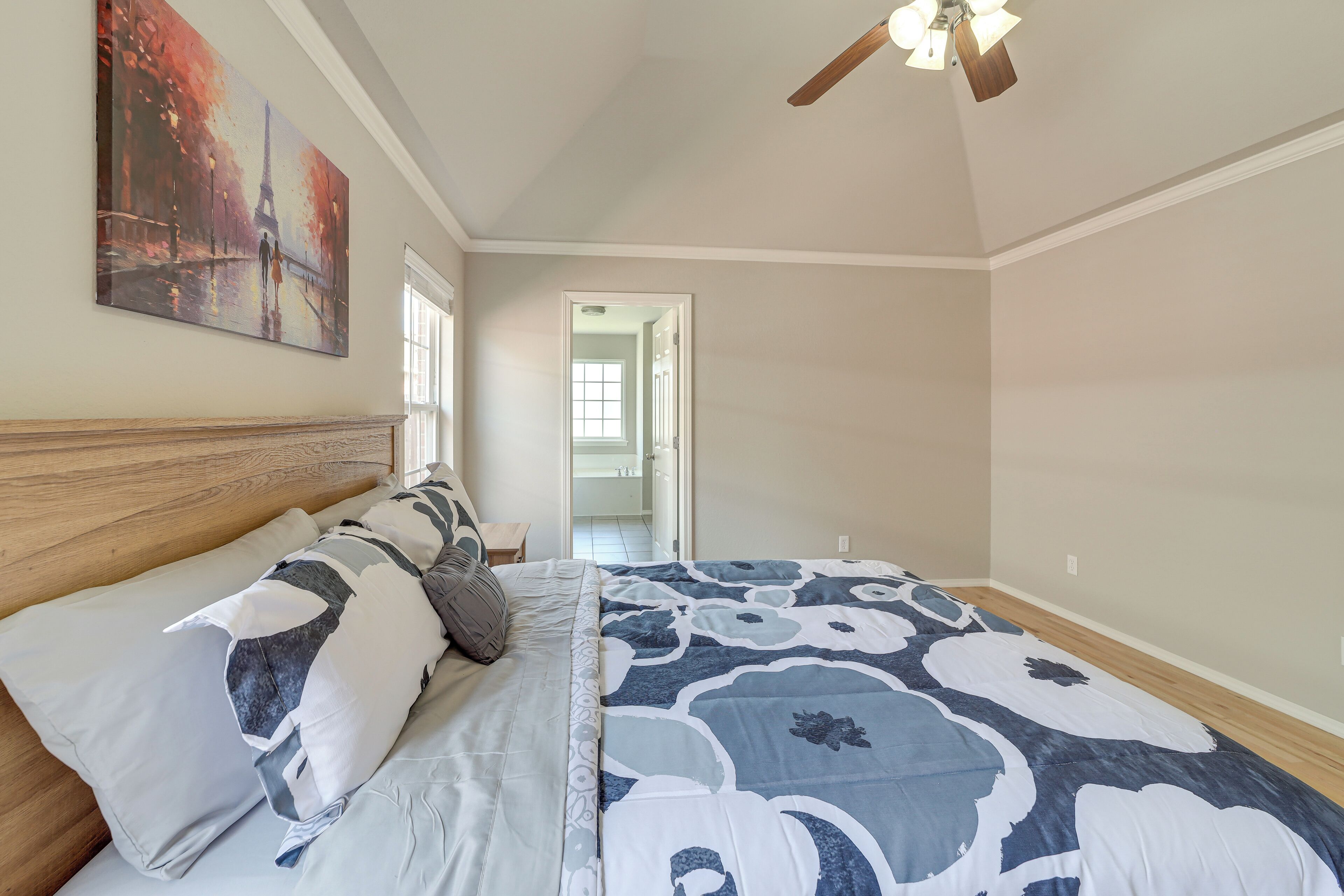 Un dormitorio con cama, ventilador de techo, un cuadro y una puerta que da a otra habitación.