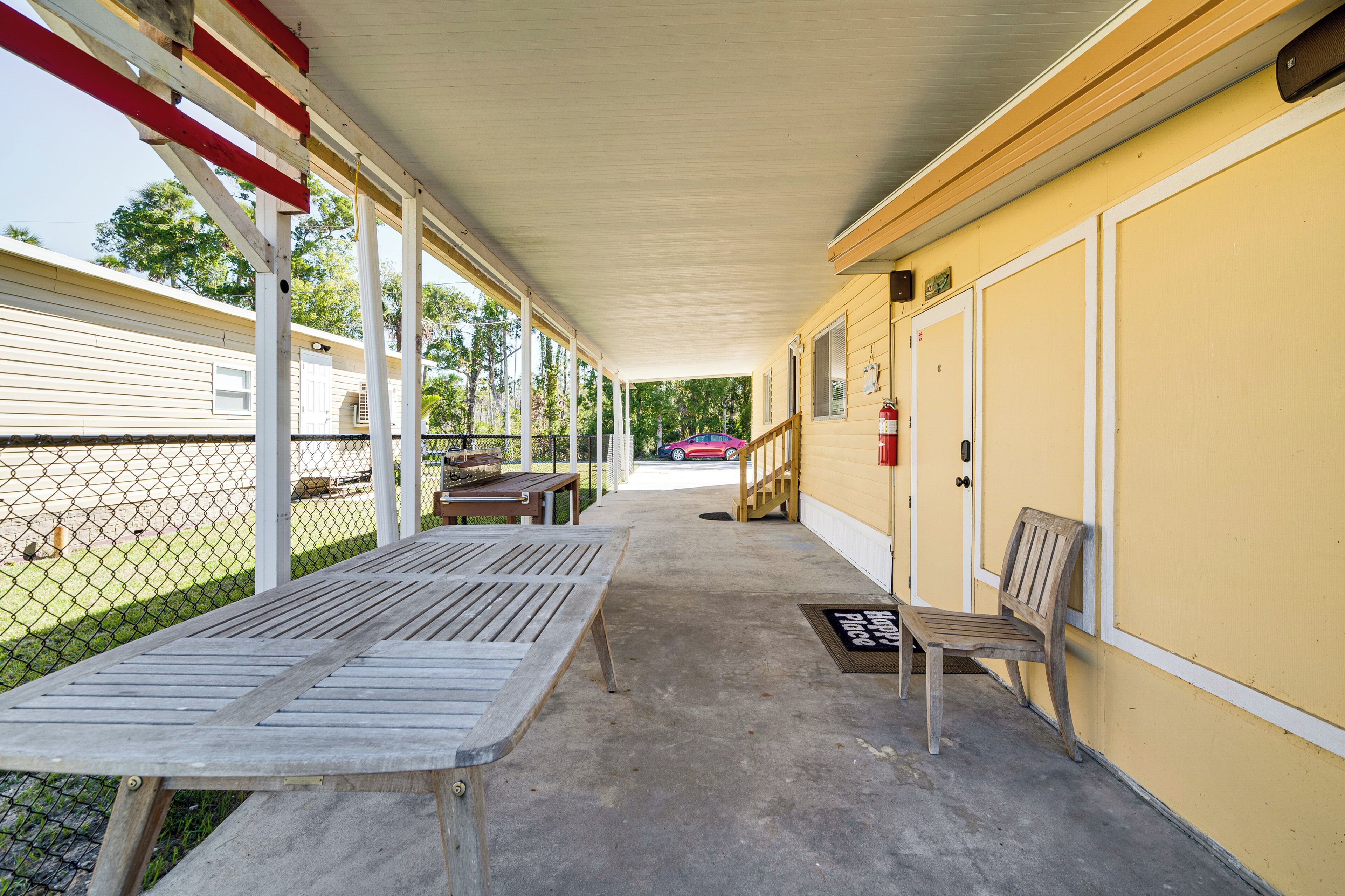 A covered outdoor area with a concrete floor, a wooden bench, a picnic table, and a yellow building with a door and window.