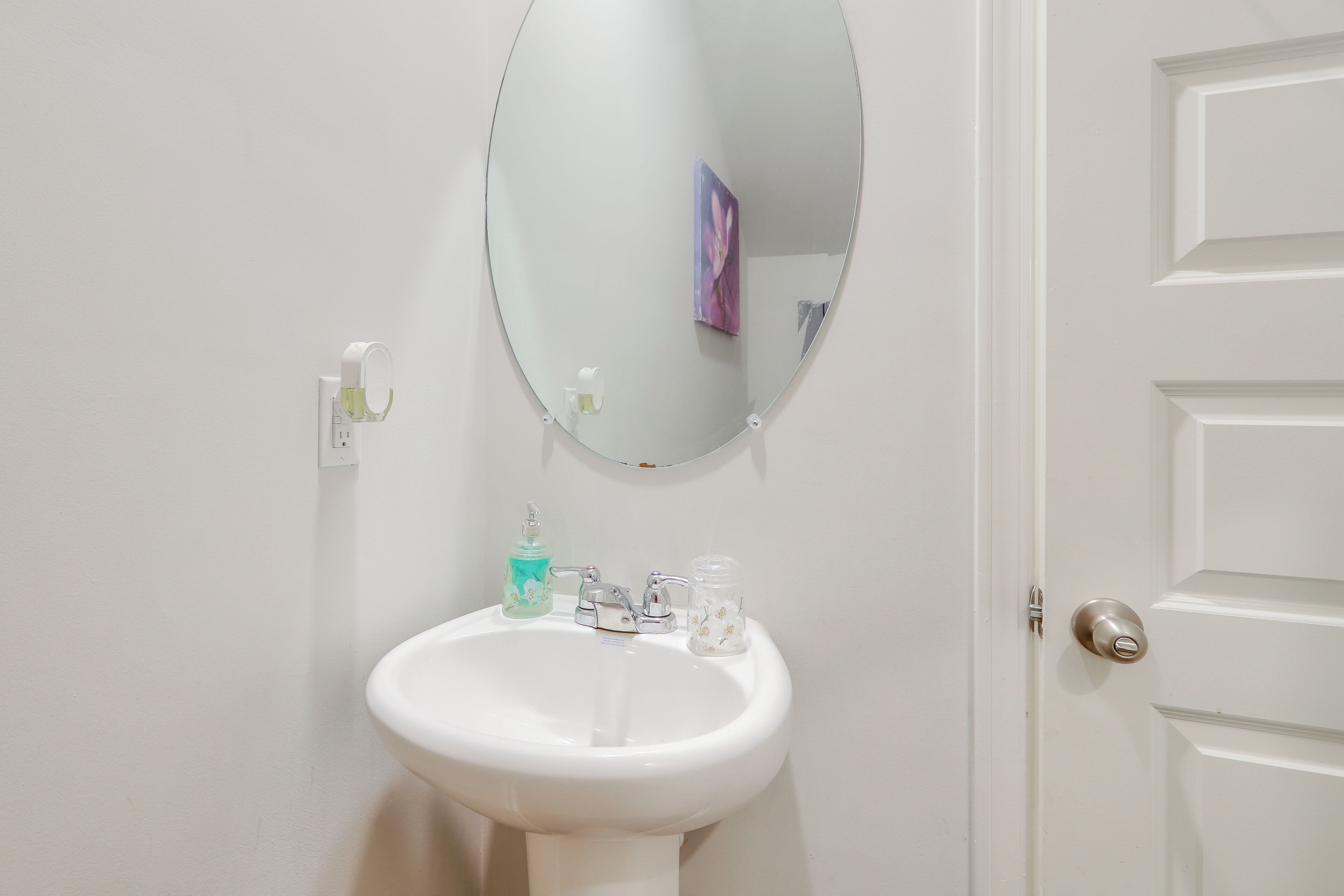 Une salle de bain blanche avec un miroir rond, un lavabo blanc et une porte dotée d’une poignée argentée.
