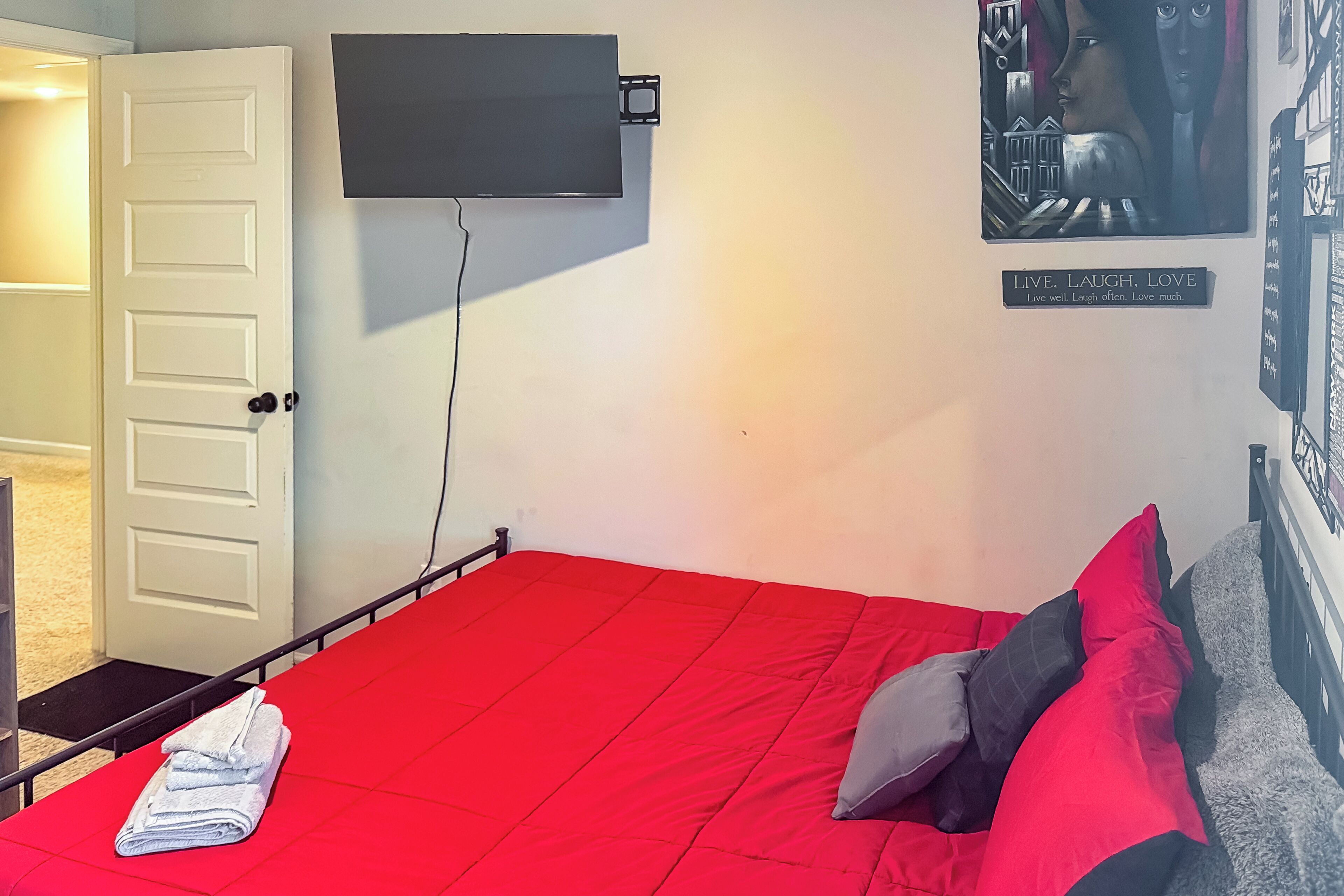 Una habitación con una colcha roja, un televisor de pantalla plana montado en la pared, una puerta y un cartel motivacional.