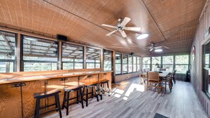 A spacious indoor area with a wooden bar, multiple stools, ceiling fans, and large windows providing natural light.