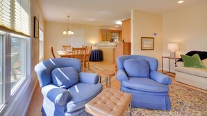 Un salon avec deux fauteuils bleus, un pouf beige, un coin repas avec une table et des chaises, une cuisine en arrière-plan et un canapé agrémenté d’un coussin vert.