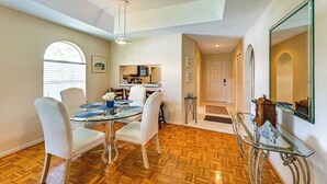 Un comedor con una mesa de vidrio, sillas blancas y piso de madera. Se ve una cocina al fondo, con electrodomésticos y una ventana con persianas.