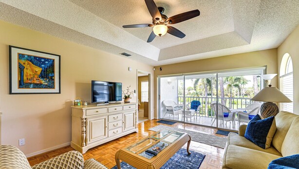 Una sala de estar con ventilador de techo, televisor, sofá, mesa de centro y un ventanal con vistas a palmeras.