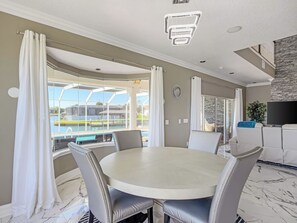 A dining area with a round table, chairs, a view of a pool, a TV, and a stone wall. - Serene Luxury Waters- Pool and canal front home in Palm Coast! (Palm Coast)