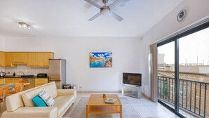 Een ruime woonkamer met een bank, salontafel en televisie. Er is een keuken met kasten en een koelkast. Aan de muur hangt een schilderij, en er is een balkon met uitzicht.