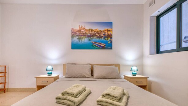 Een opgemaakt bed met twee gevouwen handdoeken, een schilderij van een haven boven het bed, en twee bedlampen op houten nachtkastjes.