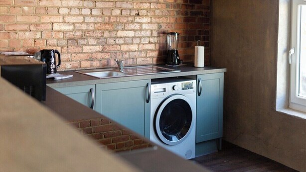 A compact kitchen with a brick wall, a washing machine, a coffee maker, and a sink. - Luxury 2 Bedroom & 2 Bathroom Central Flat (Swansea)