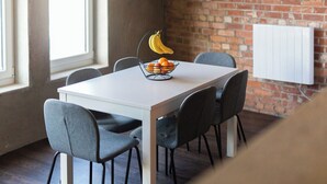A dining area with a white table, grey chairs, a fruit bowl with bananas and oranges, and a brick wall with a radiator. - Luxury 2 Bedroom & 2 Bathroom Central Flat (Swansea)