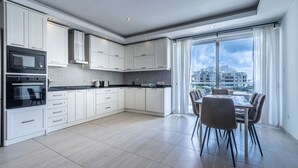 A modern kitchen with white cabinets, a black countertop, and a dining area with a table and chairs. - The Fives Apartment - St Julians (St. Julian`s)