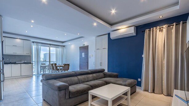 A modern living room with a grey sectional sofa, a white coffee table, and a dining area with a table and chairs. - The Fives Apartment - St Julians (St. Julian`s)