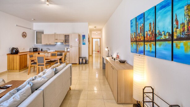 A modern apartment with a living room, dining area, and kitchen. There is a sofa, a dining table, and a kitchen counter. The apartment has a cityscape painting on the wall. - The Beach Apartment - Sliema (Sliema)