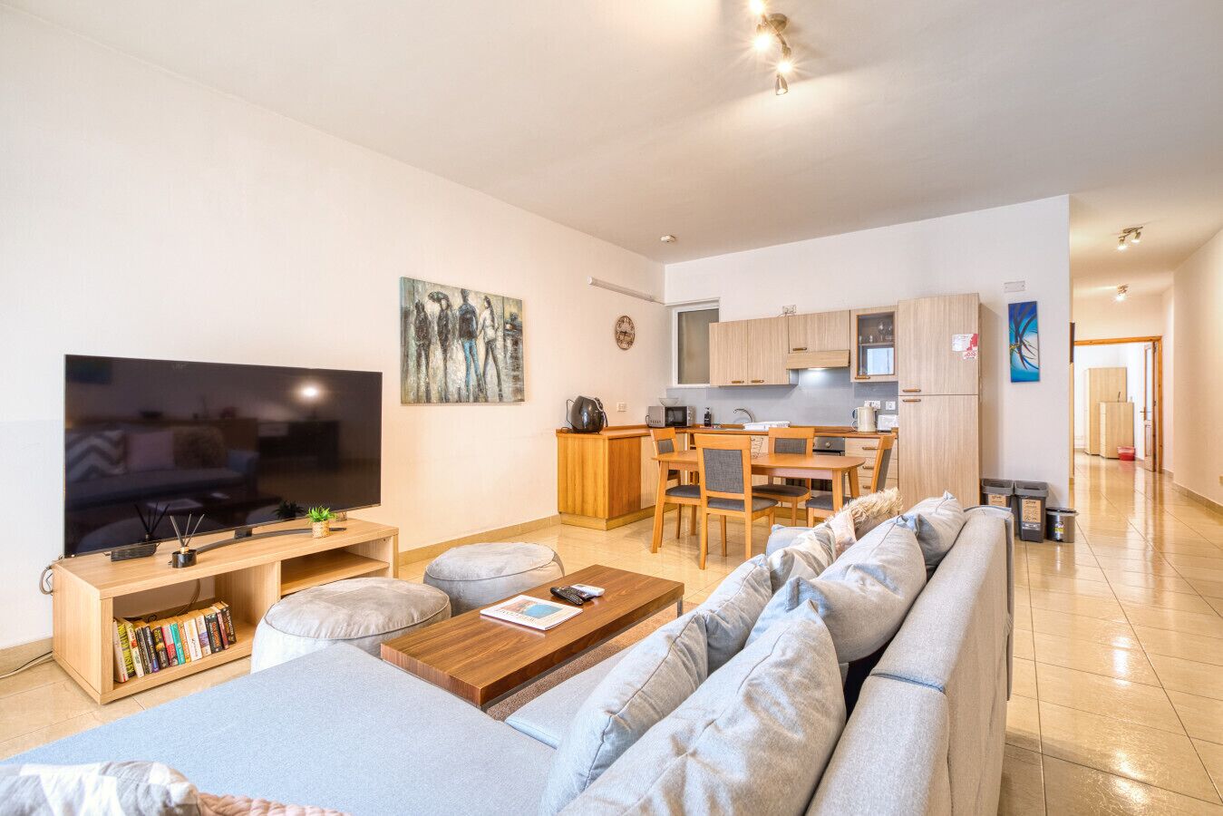 Una sala moderna con un televisor de pantalla plana grande, una mesa de centro de madera y un sofá gris. Hay una zona de cocina con armarios de madera y una mesa de comedor.