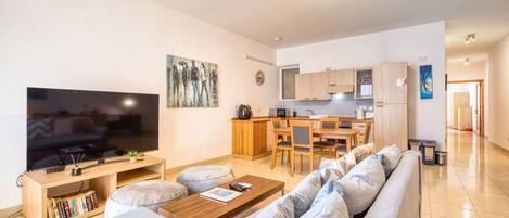 Una sala moderna con un televisor de pantalla plana grande, una mesa de centro de madera y un sofá gris. Hay una zona de cocina con armarios de madera y una mesa de comedor.