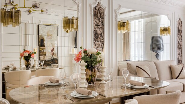 A dining area with a round table set for four, surrounded by chairs. A vase with flowers is placed on the table, and there are candles and wine glasses. The room has a classic design with a large mirror and decorative wall panels. - Merveil Luxury Suite - Louvre Aboukir IX (Paris)