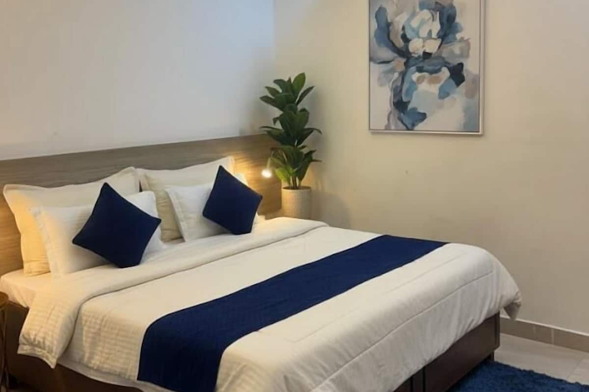 Een net opgemaakt bed met wit- en blauw beddengoed, een blauw vloerkleed en een wandairconditioner.
