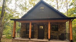 Une cabane en pierre avec un toit sombre, une véranda à l’avant soutenue par des piliers en bois et une porte surmontée d’une fenêtre.