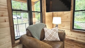 L'intérieur d'un chalet en bois est aménagé avec un canapé en cuir marron, un téléviseur fixé au mur et de grandes fenêtres qui offrent une vue sur l'extérieur.