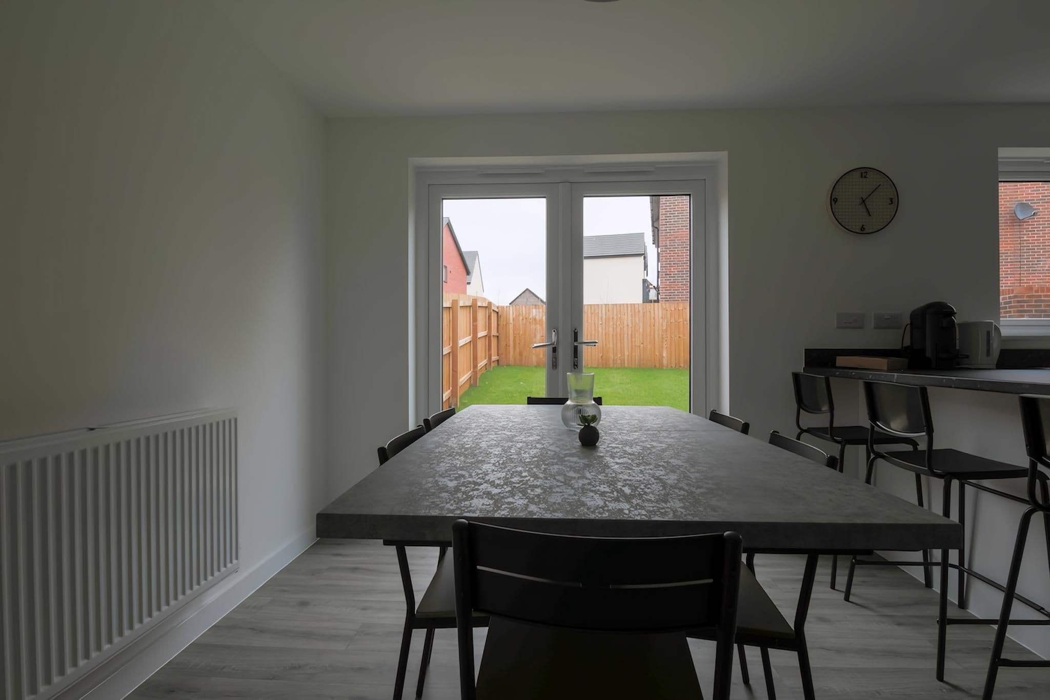 A dining area with a table and chairs, a window with a clock, and a view of a backyard.