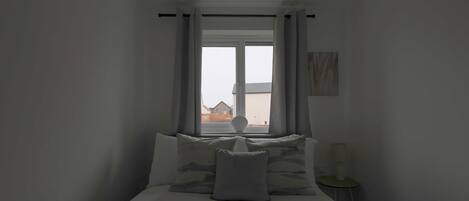 A bedroom with a bed, pillows, a bedside table, a lamp, and a window with curtains.