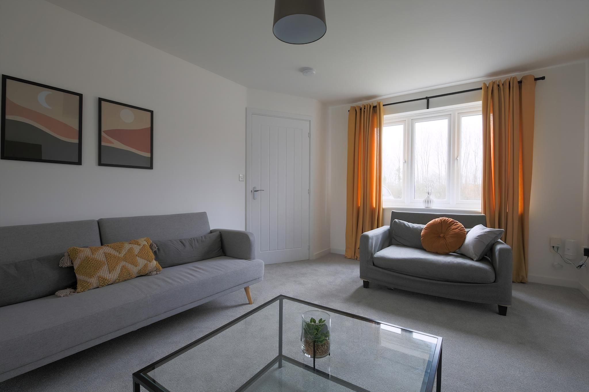 A living room with a grey sofa, a glass coffee table, and a window with curtains.