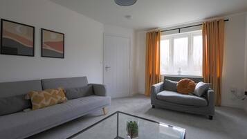 A living room with a grey sofa, a glass coffee table, and a window with curtains.