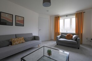 A living room with a grey sofa, a glass coffee table, and a window with curtains. - The Haven Modern 4-bed Home With Garden (Cardiff)