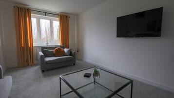Una sala de estar moderna con un sofá, una mesa de centro de vidrio, un televisor de pantalla plana y una ventana con cortinas.