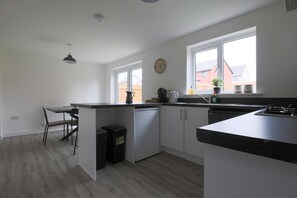 Uma cozinha moderna com armários brancos, bancada preta, espaço de refeições com uma cadeira e janela com vista para edifícios.