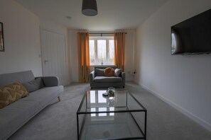 A living room with a sofa, a glass coffee table, a TV mounted on the wall, and a window with curtains.