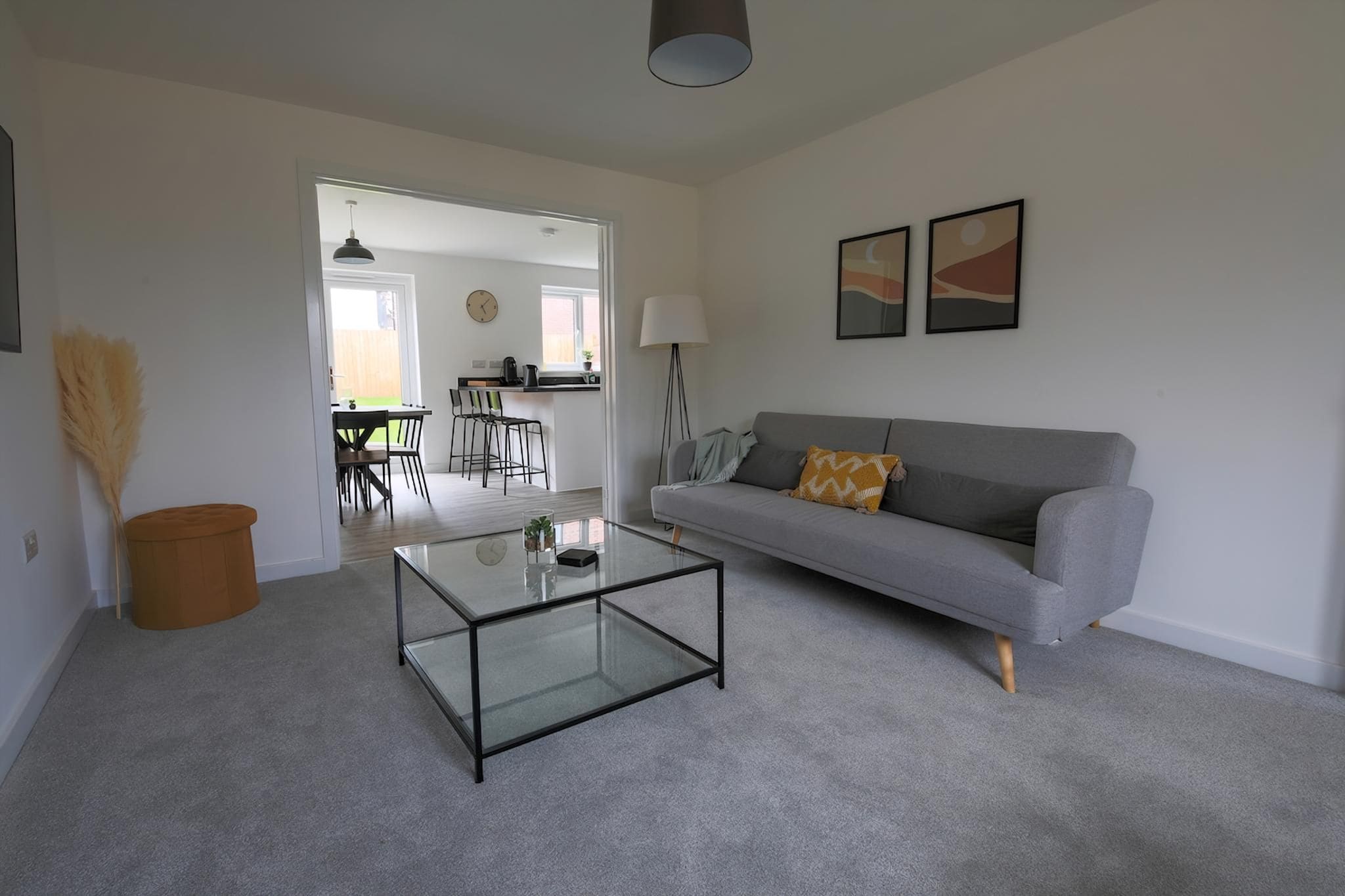 A living room with a glass coffee table, a grey sofa with a patterned cushion, a lamp, and two wall-mounted pictures.